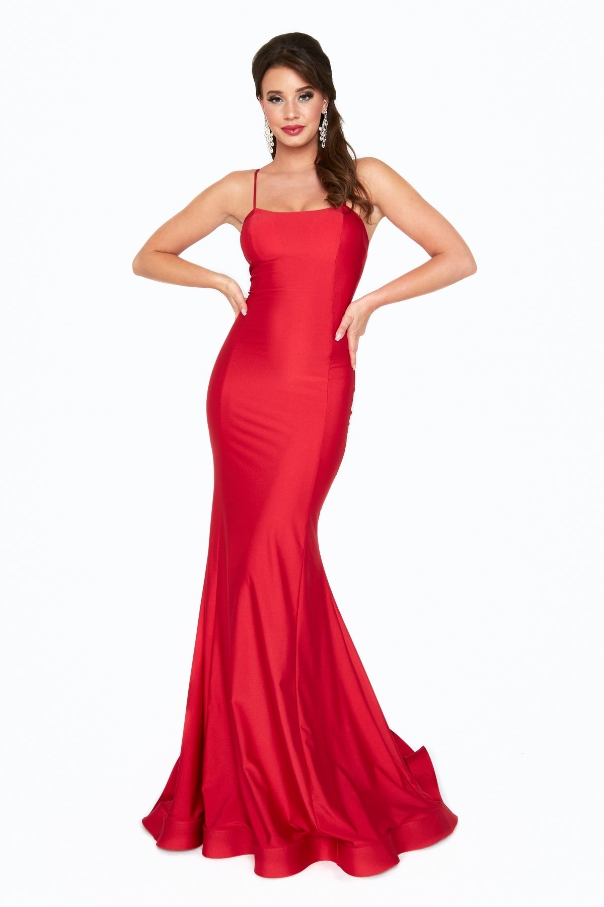 Woman wearing the Atria 6520H Lace-Up Back Long Prom Dress—a fitted, floor-length red gown with thin straps—stands with hands on hips against a plain white background.
